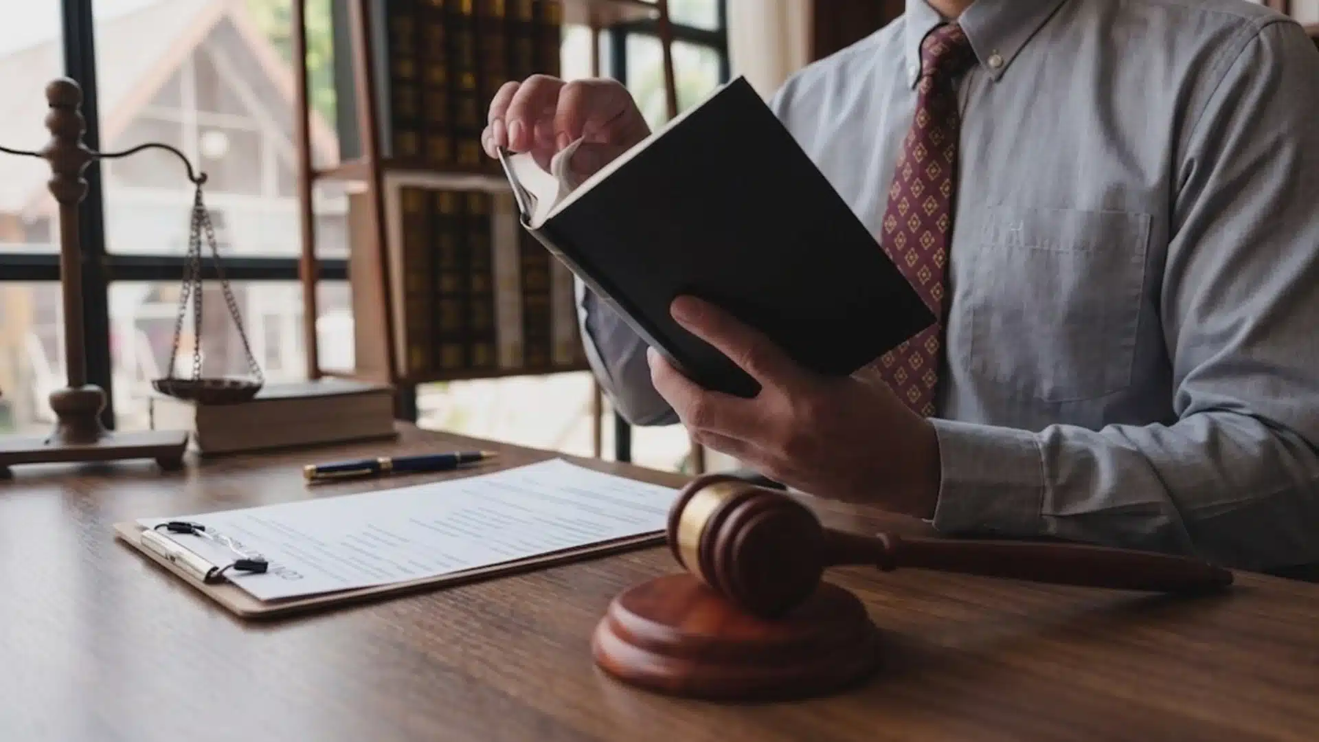 A lawyer in a light shirt and patterned tie reads a book at a dark wooden desk with a gavel and scales of justice