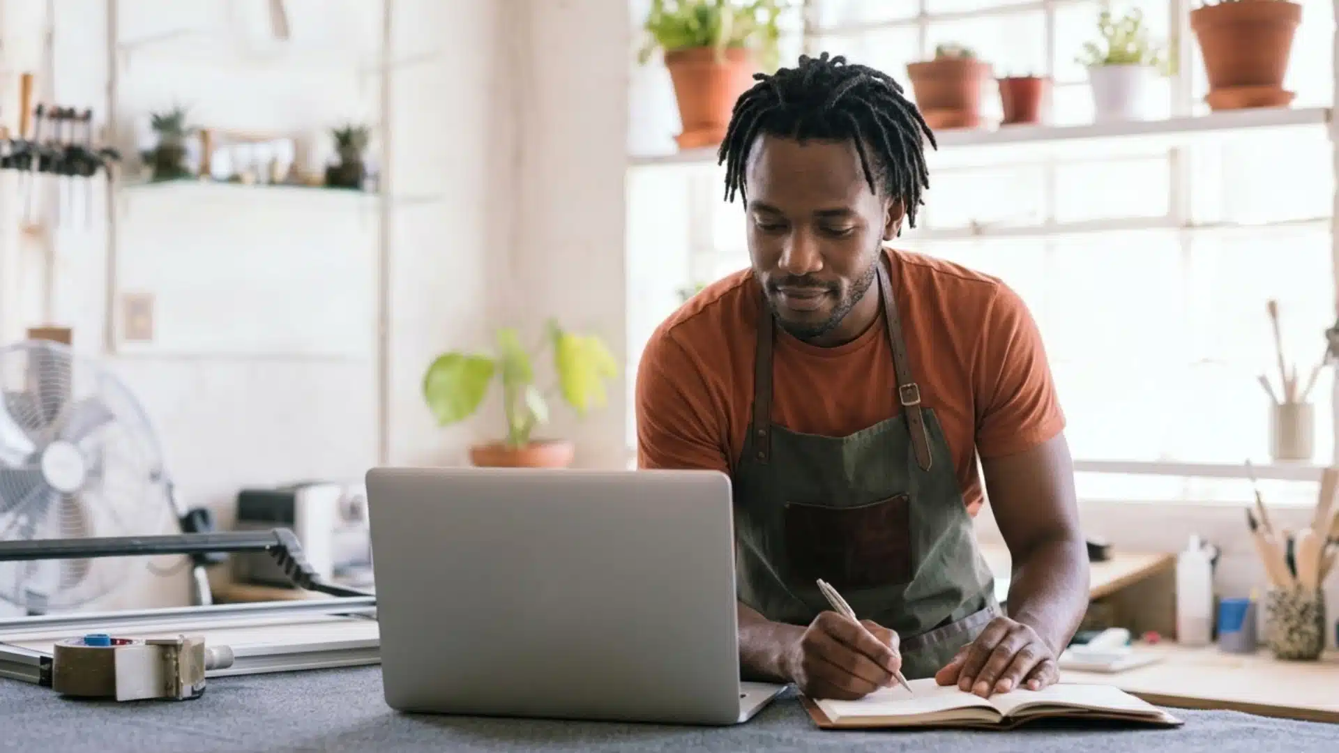 A man with dreadlocks wearing an apron writes in a notebook behind a laptop in a bright, plant-filled workshop