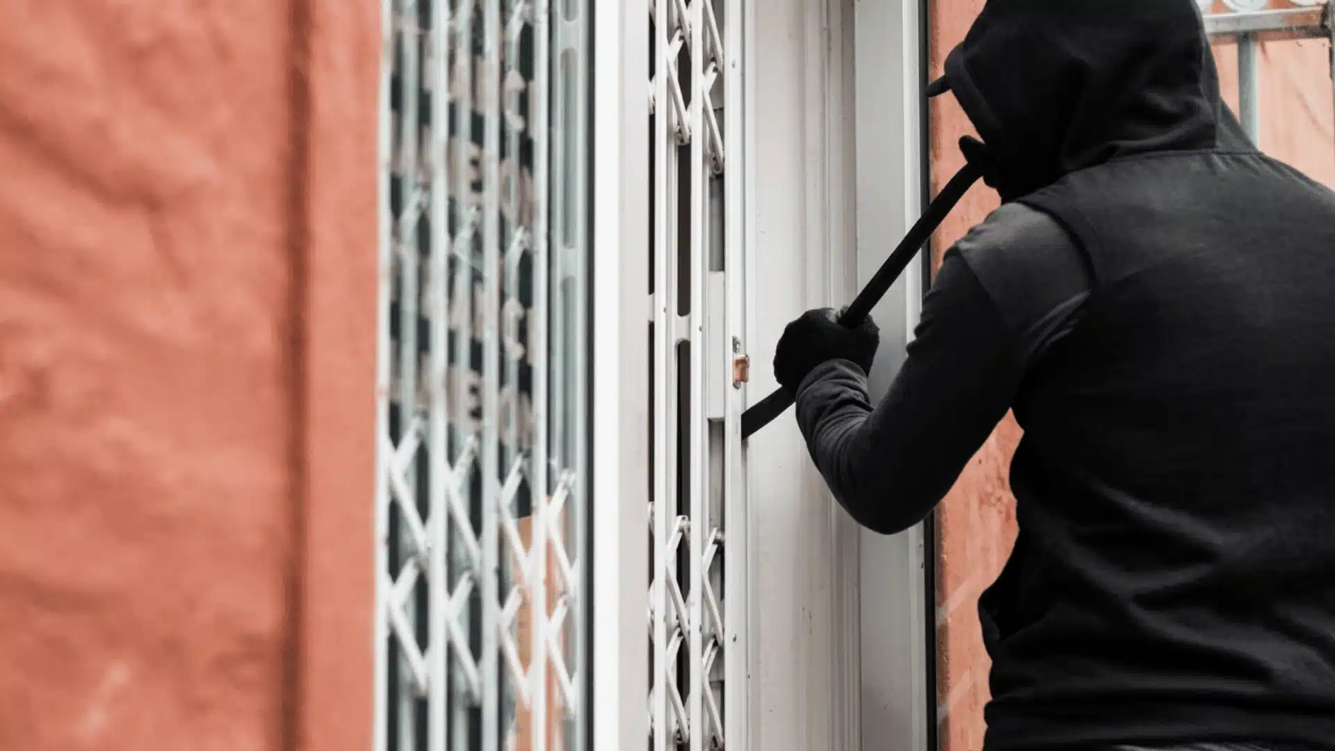 A person in a black hoodie pries open a white security gate with a crowbar next to an orange wall