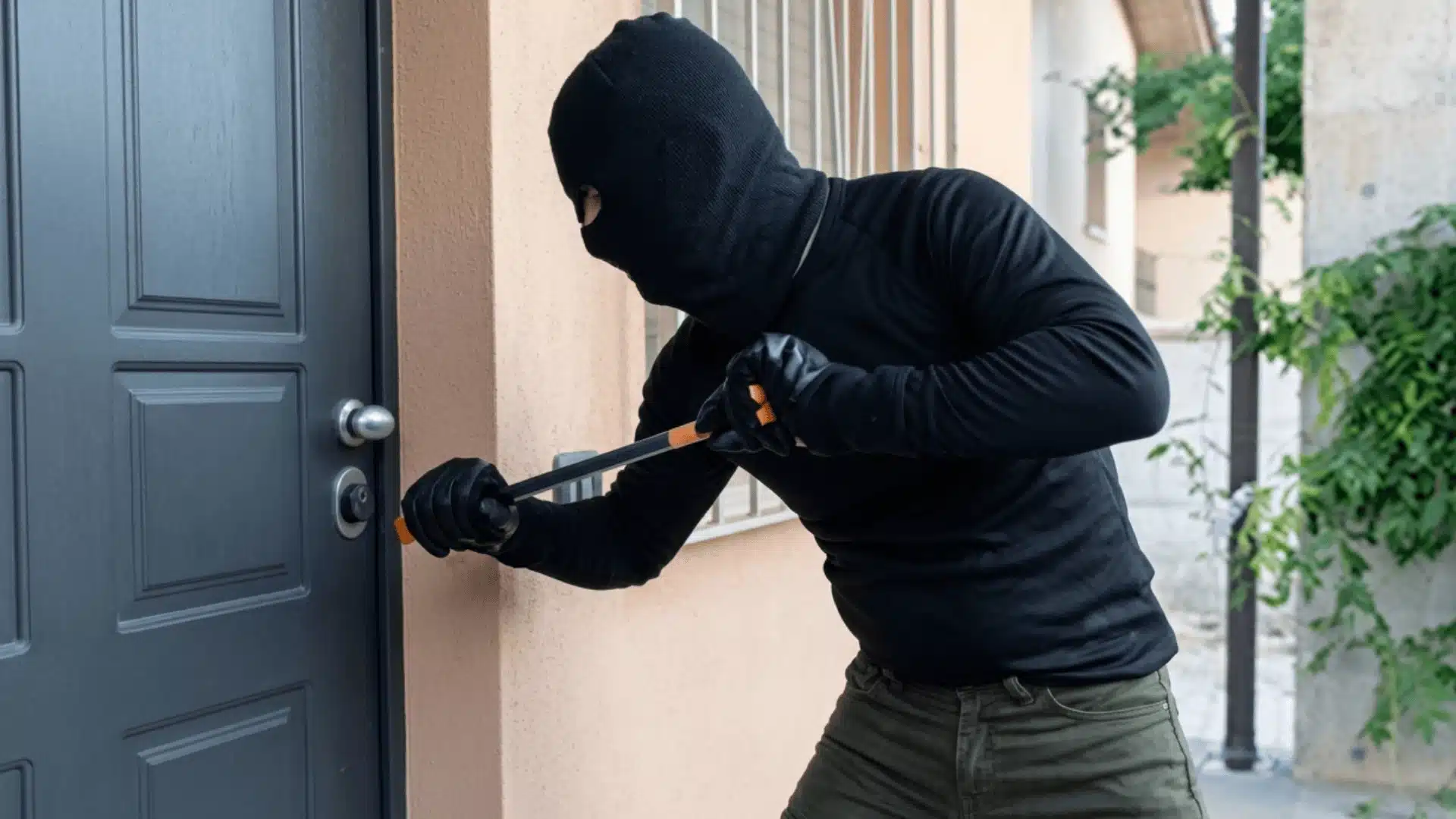 A person in a black ski mask uses a crowbar to pry open the lock of a dark grey front door