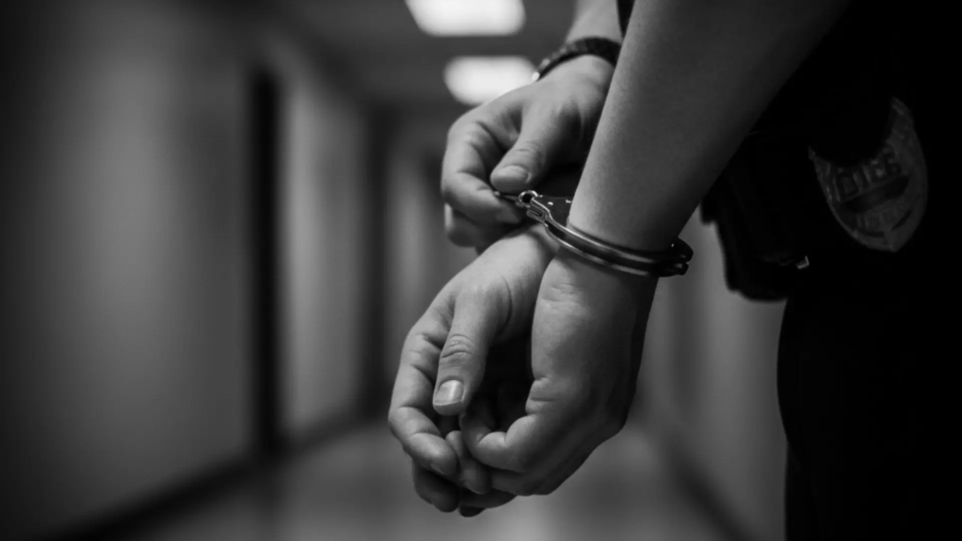 A person's hands are shown in handcuffs, held by a police officer in a dimly lit hallway. The black and white image conveys a serious tone