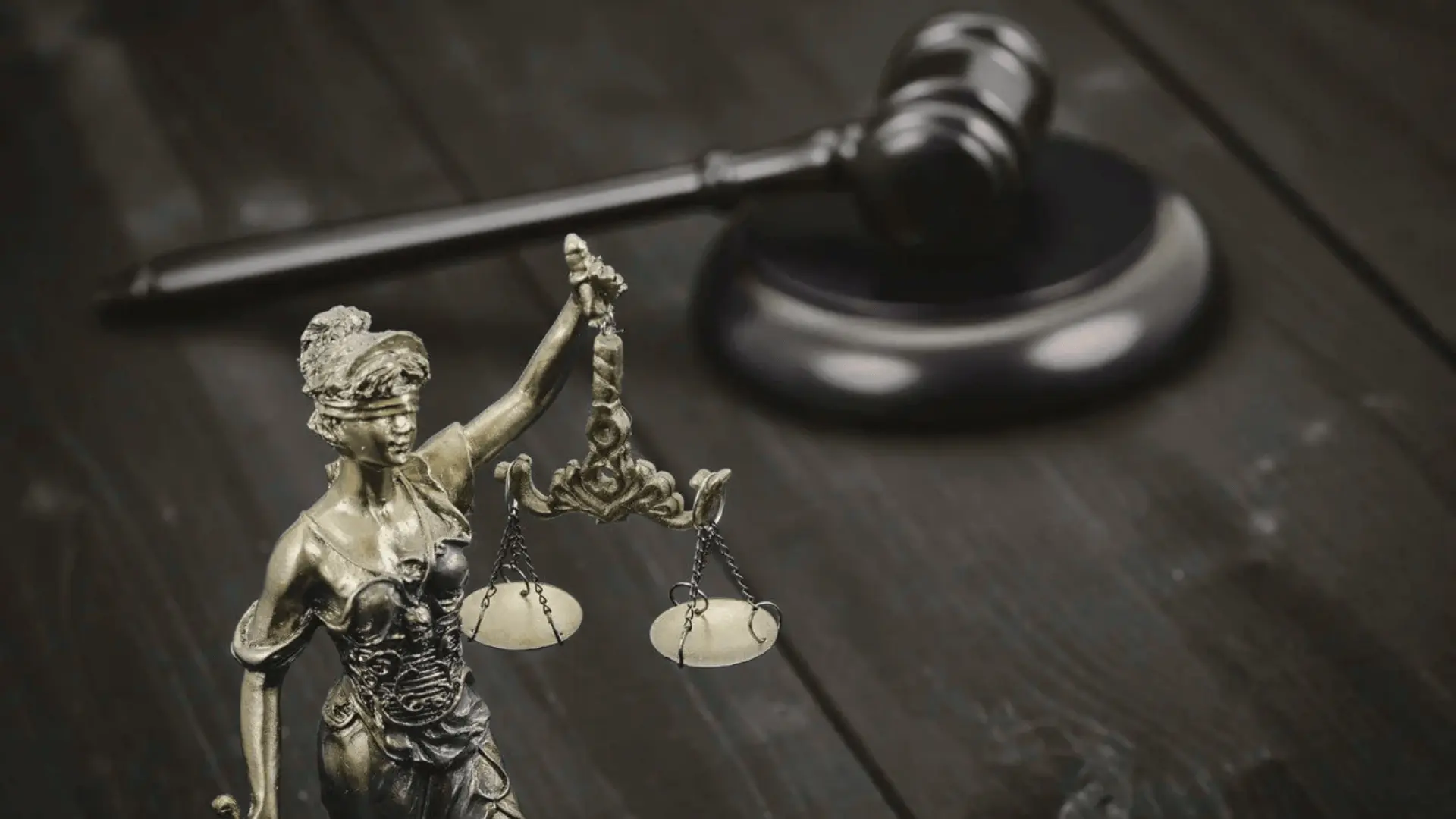 Bronze Lady Justice statue holding scales stands on a dark wooden table, with a blurred gavel in the background, symbolizing law and fairness
