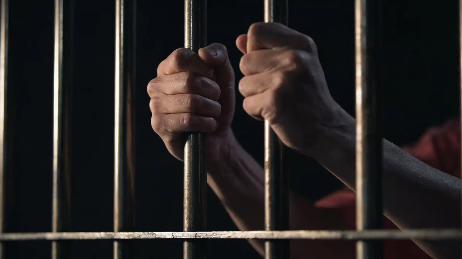 Close-up of hands gripping metal prison bars inside a dark jail cell, symbolizing confinement