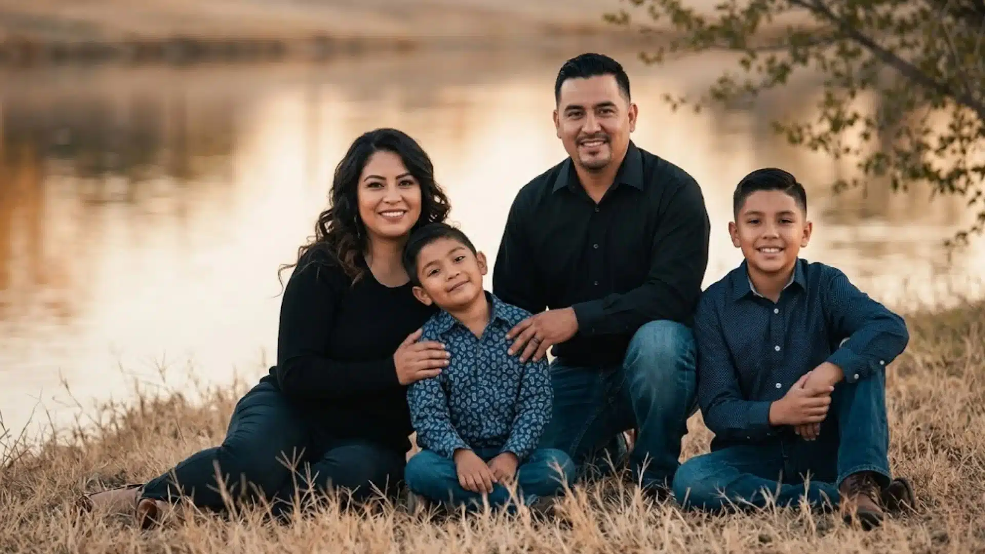 Family of four sitting by a lakeside at sunset, smiling together in a relaxed outdoor portrait setting