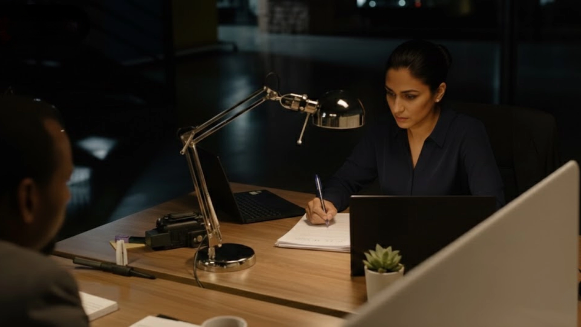Female private investigator reviewing documents at desk with laptop and lamp during late night office consultation