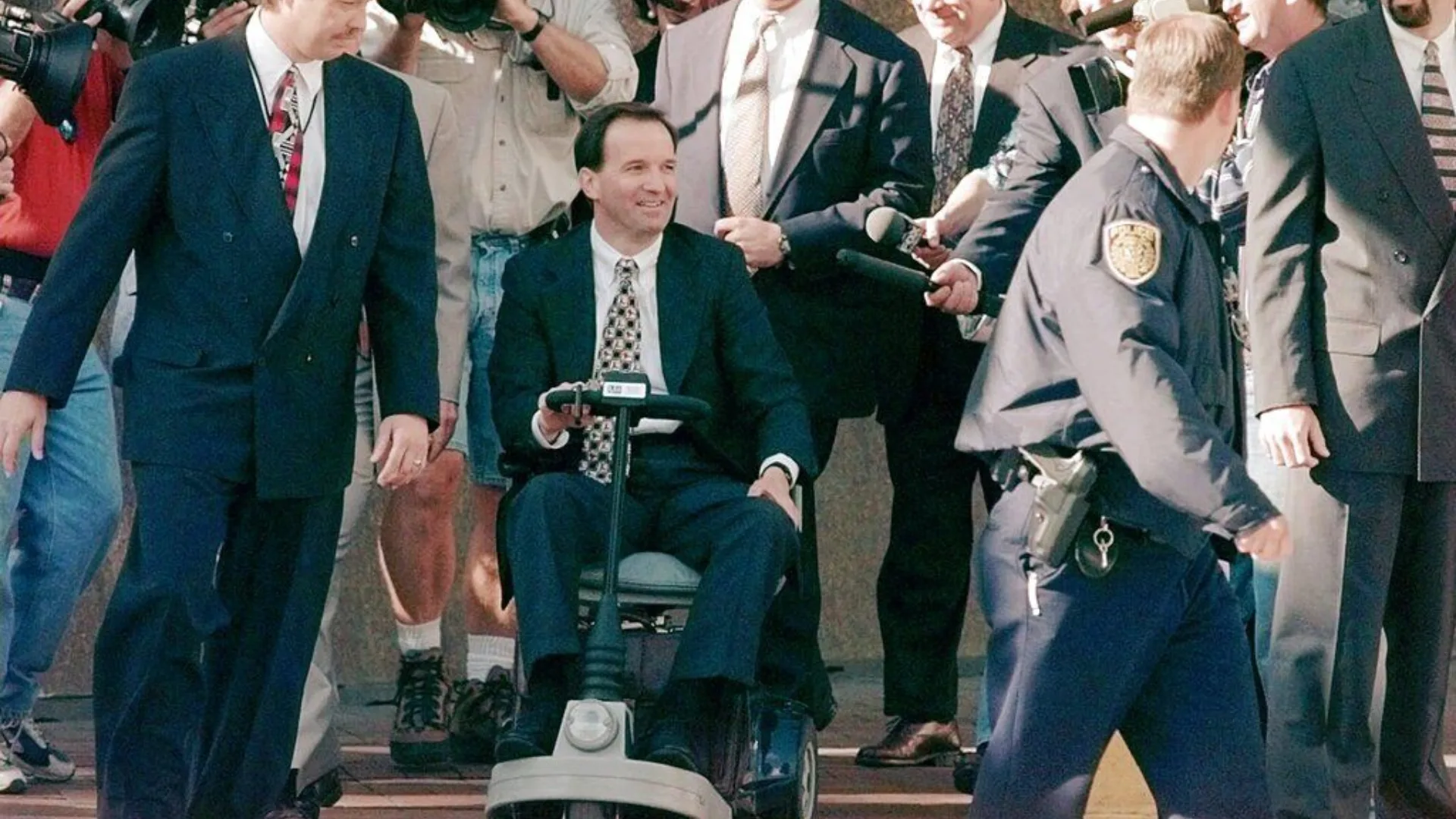 Man in suit using mobility scooter exits building surrounded by reporters, cameras, and police officers