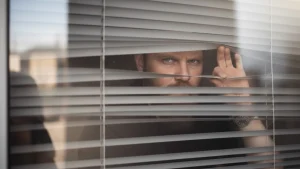 Man peeking through window blinds, secretly watching from inside, illustrating voyeurism crime and invasion of privacy concept
