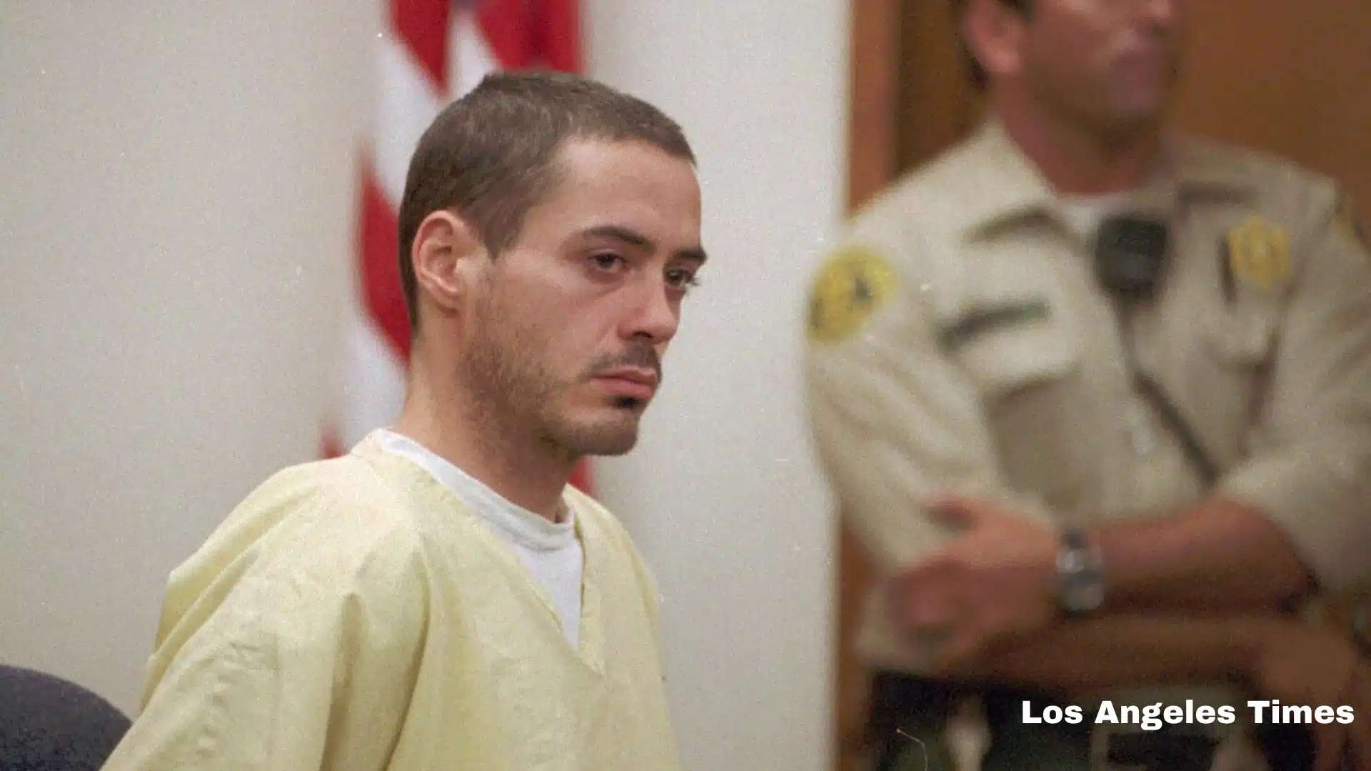Robert Downey Jr. with short hair and a yellow shirt sits in a courtroom as a sheriff stands behind him