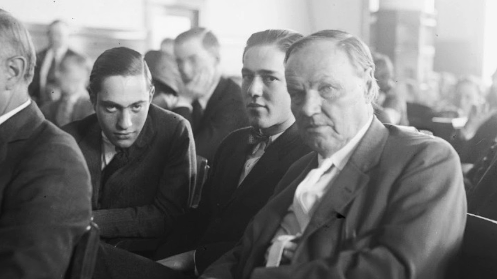 Three men seated closely in a crowded 1920s courtroom during a high profile criminal trial.
