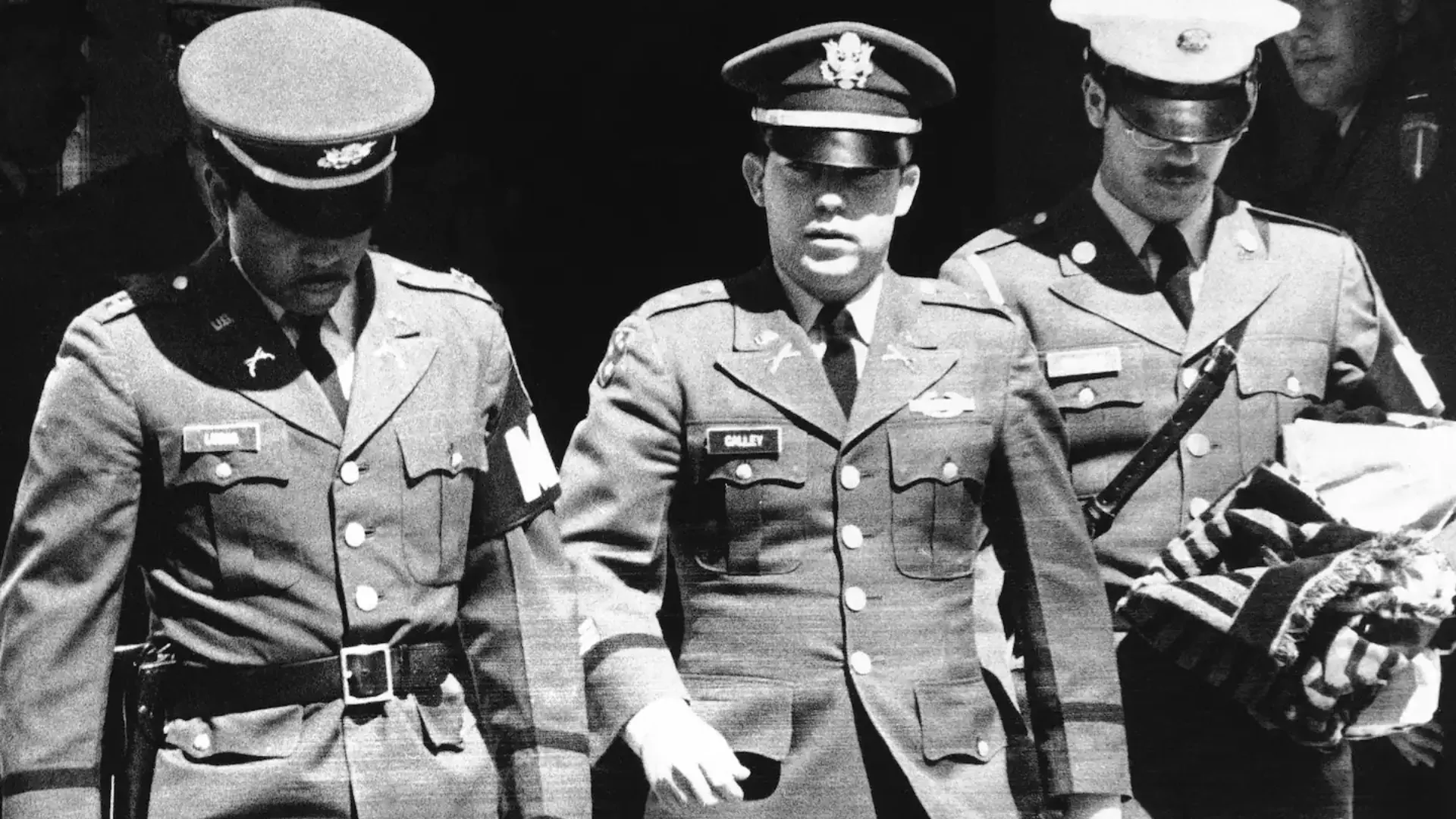 Three uniformed officers escort a man in military attire, one carrying a folded flag, in black and white scene