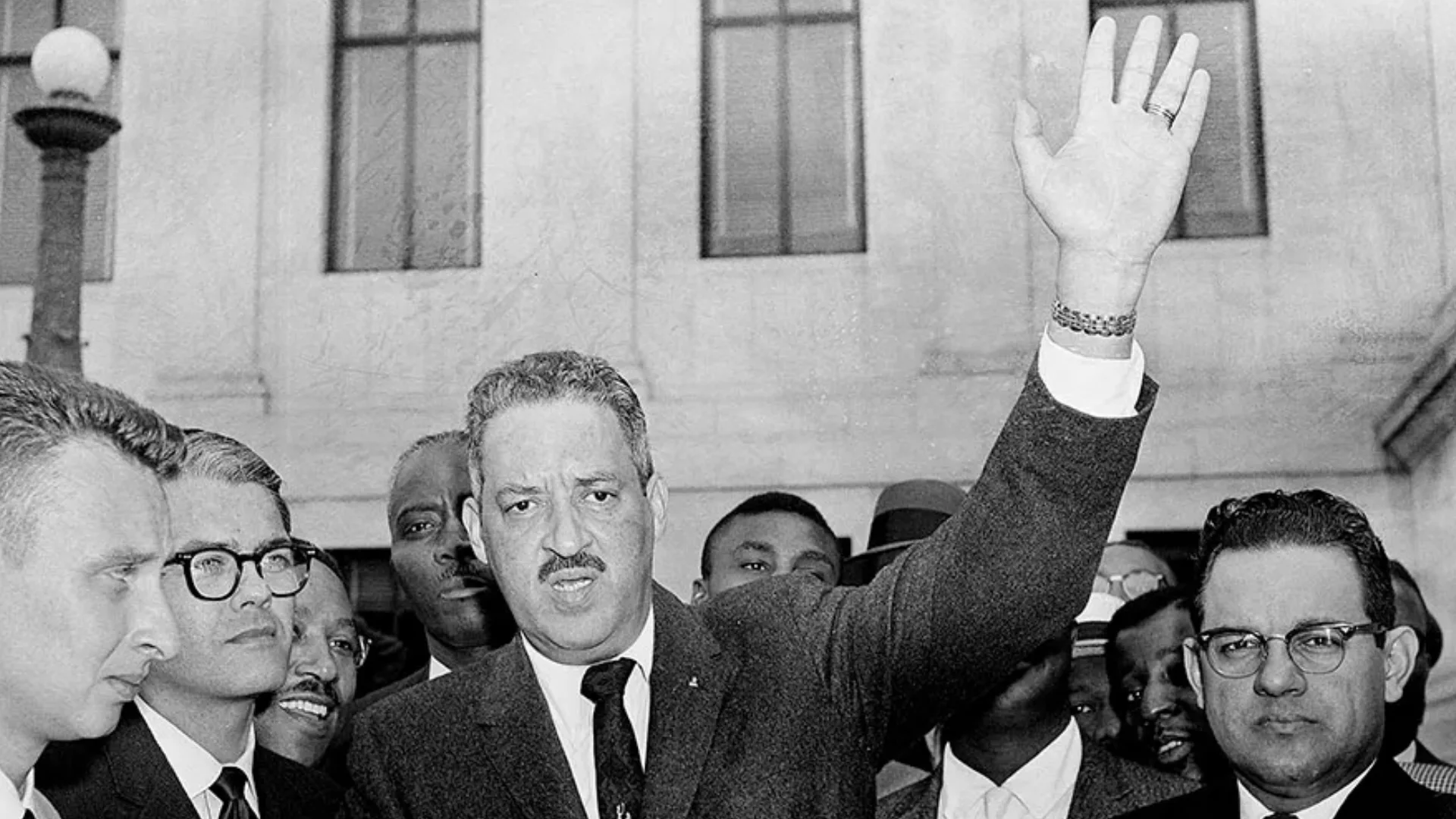 Thurgood Marshall speaking to reporters outside courthouse, raising hand during 1950s segregation case.