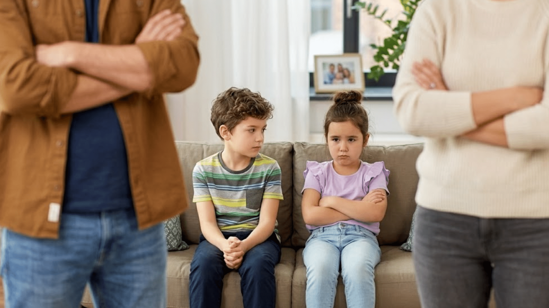 Two children sitting on couch looking upset while parents stand apart with crossed arms during family conflict at home