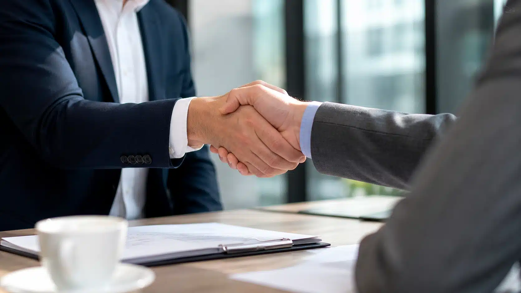 Close-up handshake between two professionals over documents in modern office with coffee cup nearby