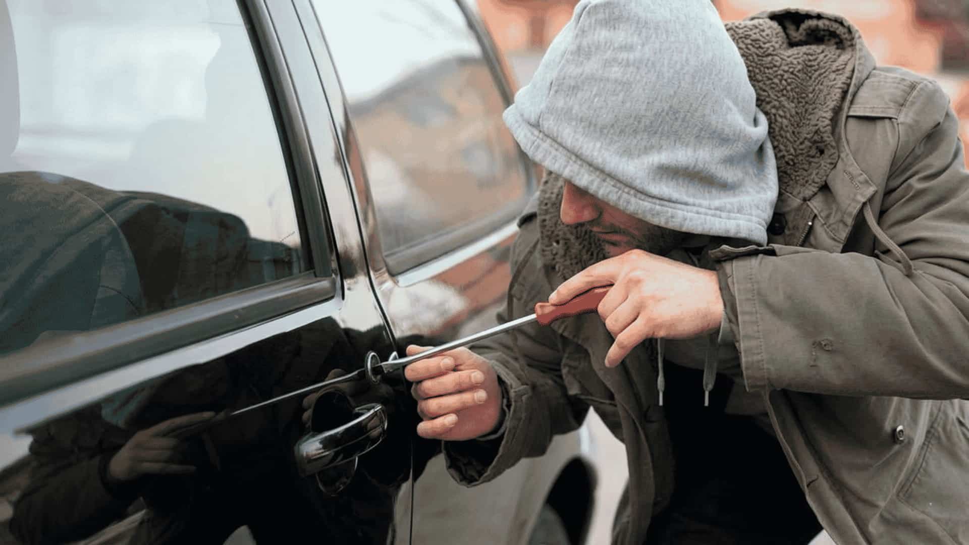A person in a hooded jacket uses a tool on a black car door handle