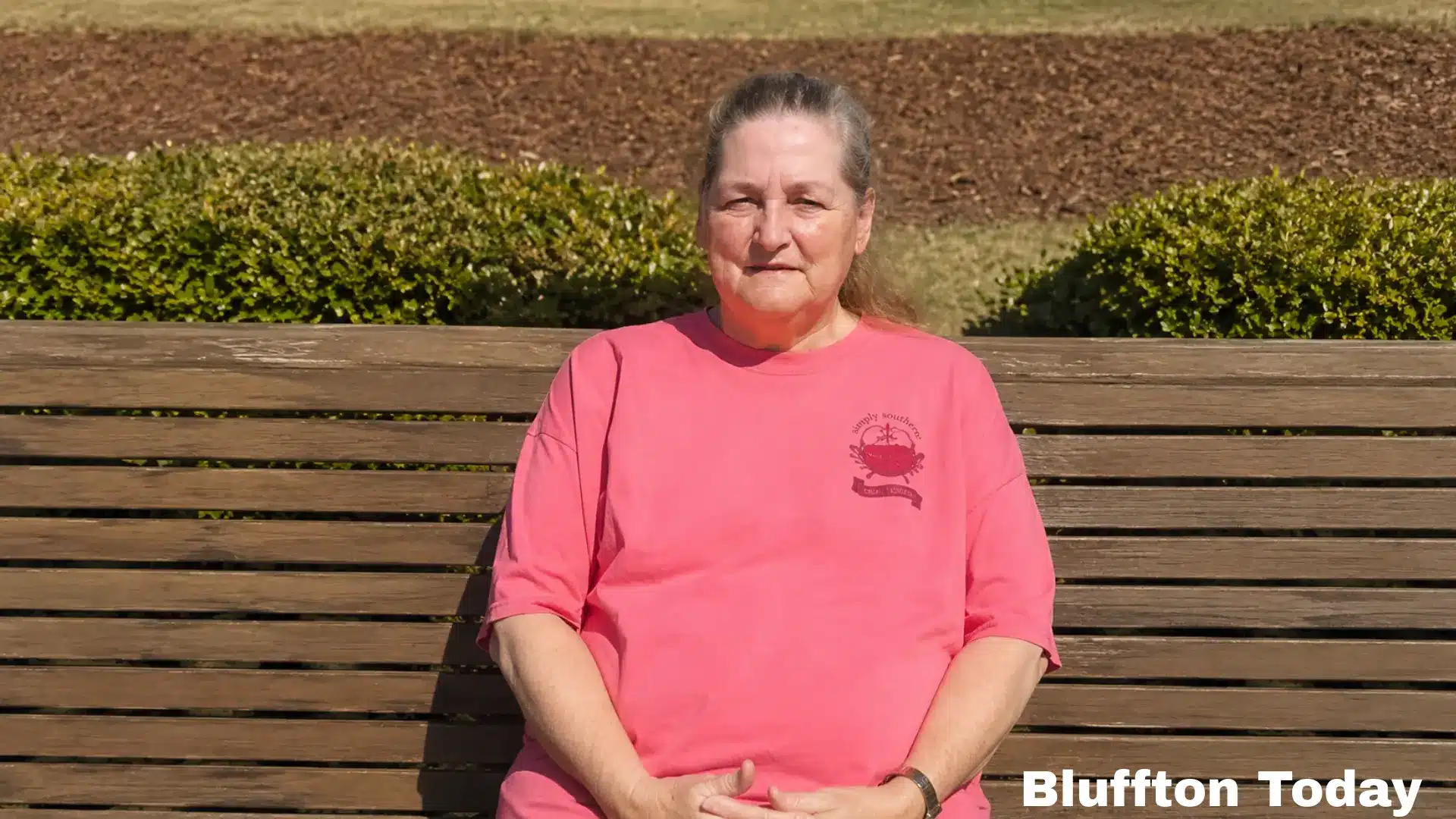 A woman in a pink t-shirt sits on a wooden park bench outdoors with green bushes and mulch in the background