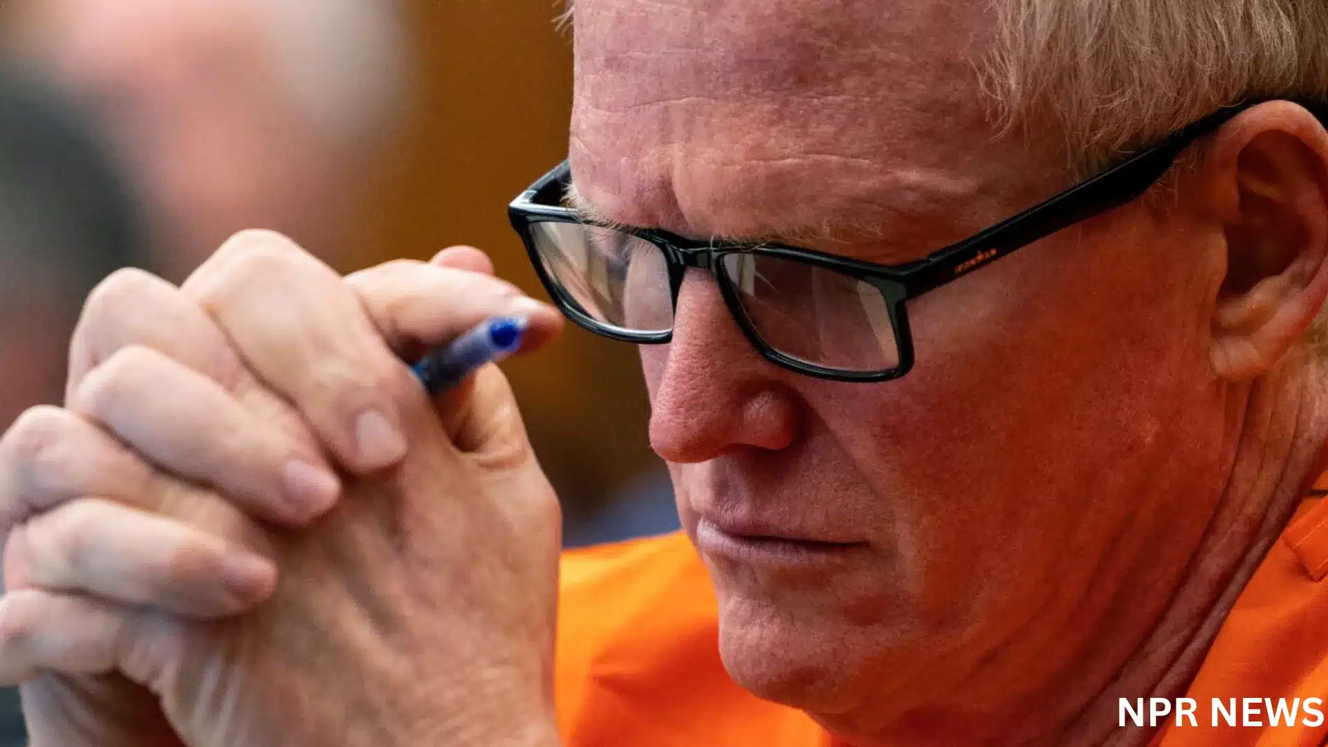 Close-up of an older man with glasses and an orange shirt, holding a pen and looking down pensively