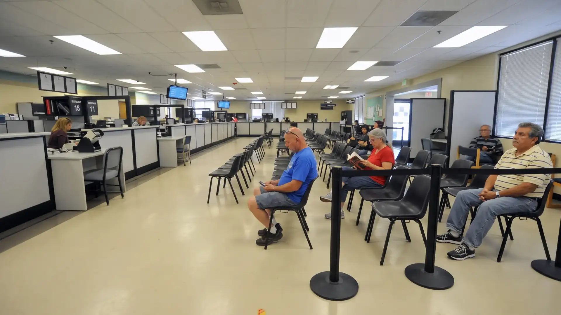 DMV waiting area with people seated in queue, service counters, and bright overhead lighting in office interior