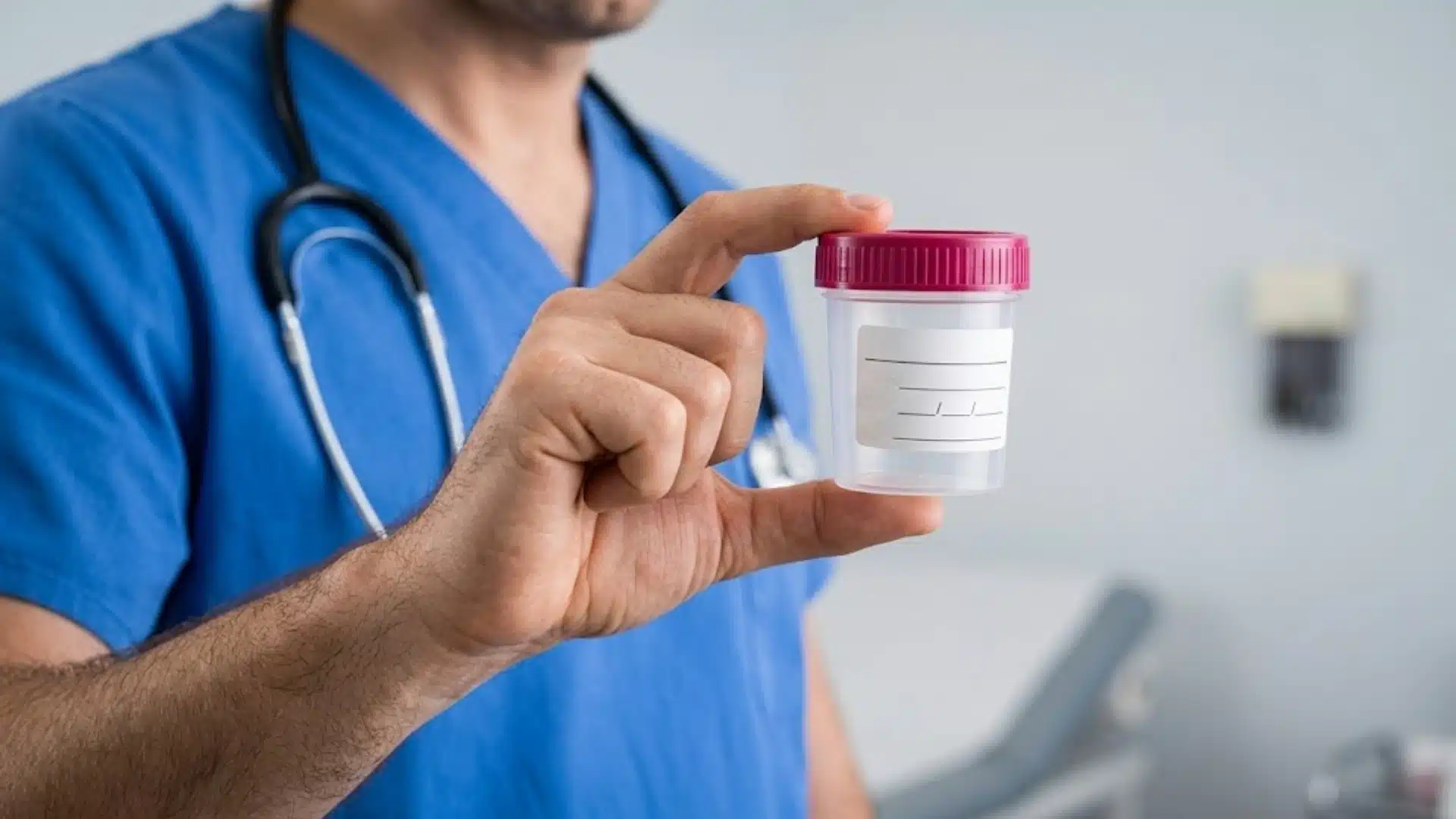 Doctor in blue scrubs holding a labeled urine sample container in a clinical exam room