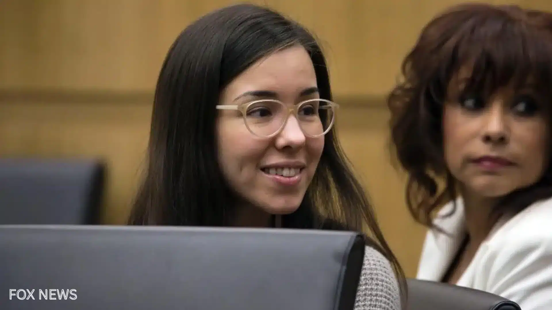 Jodi Arias sitting in court during trial proceedings, smiling while wearing glasses beside another woman in the courtroom