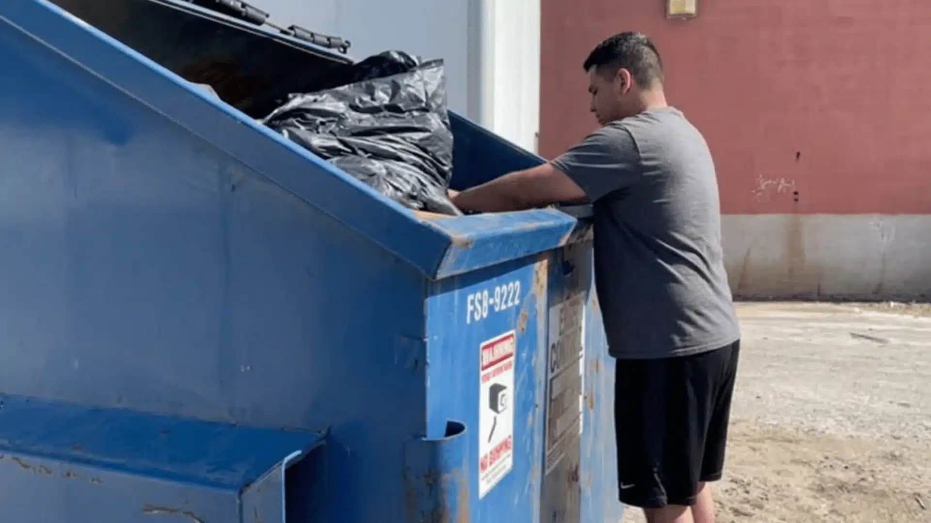 Man searching through a large blue dumpster outdoors near a commercial building during daytime