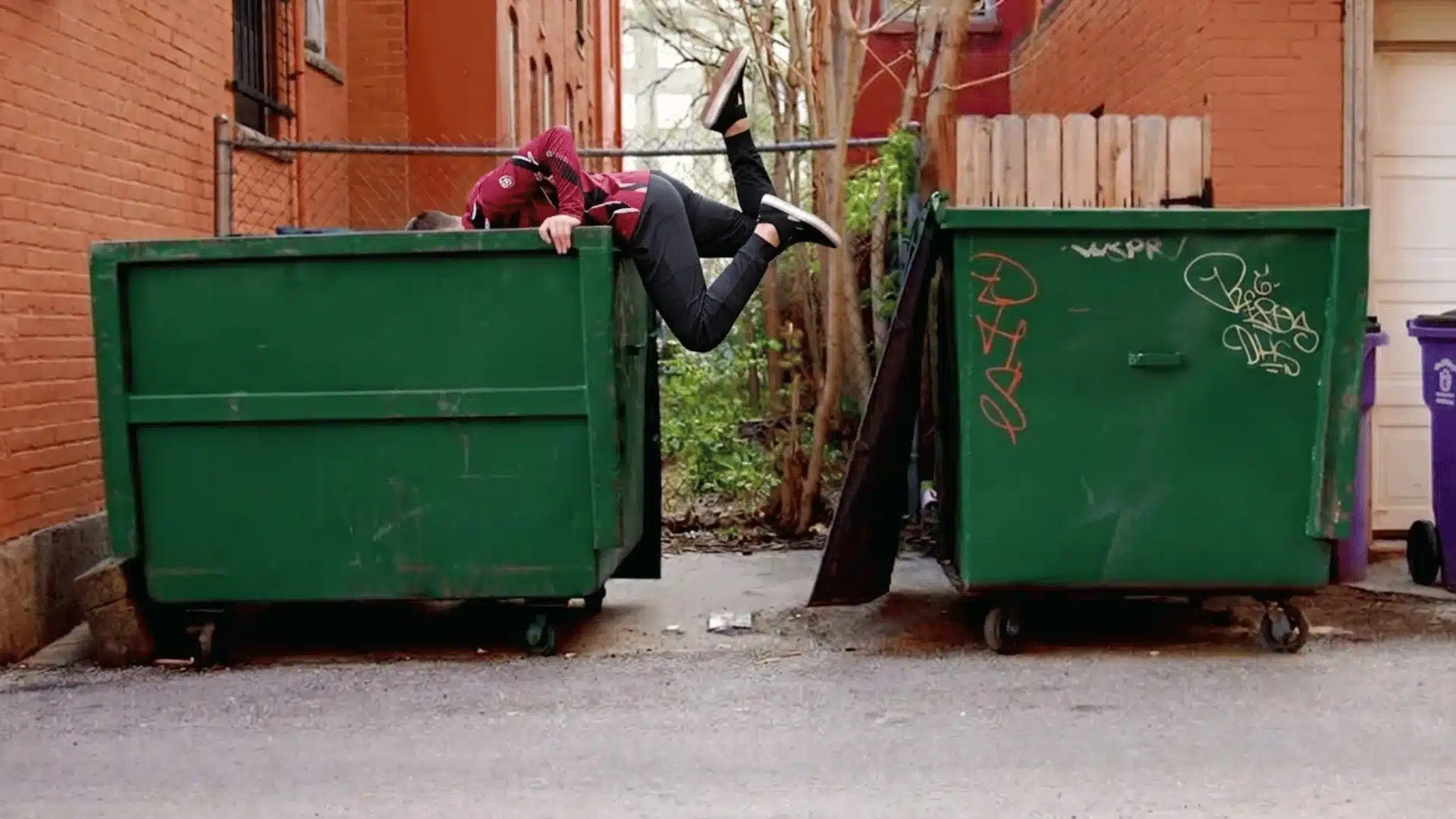 Person dumpster diving in a large green trash container behind buildings in a narrow urban alleyway
