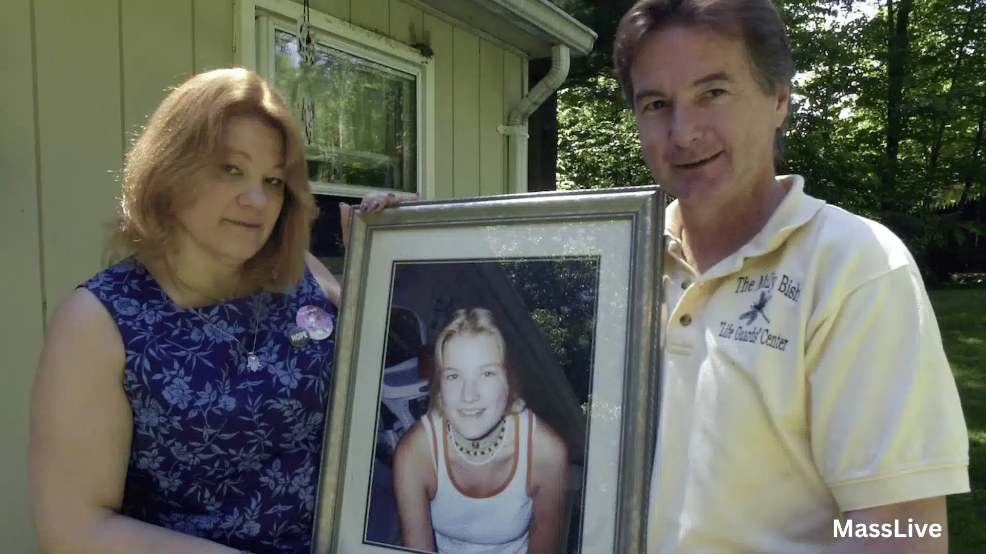 Two adults standing outdoors holding a framed portrait of a teenage girl