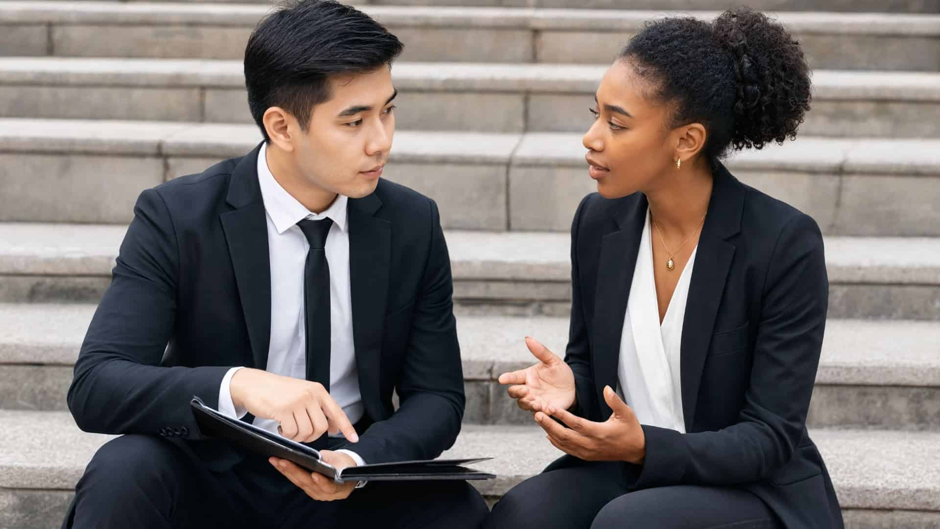 East Asian man and a Black woman in business suits sit on stone steps, discussing a document in an open folder