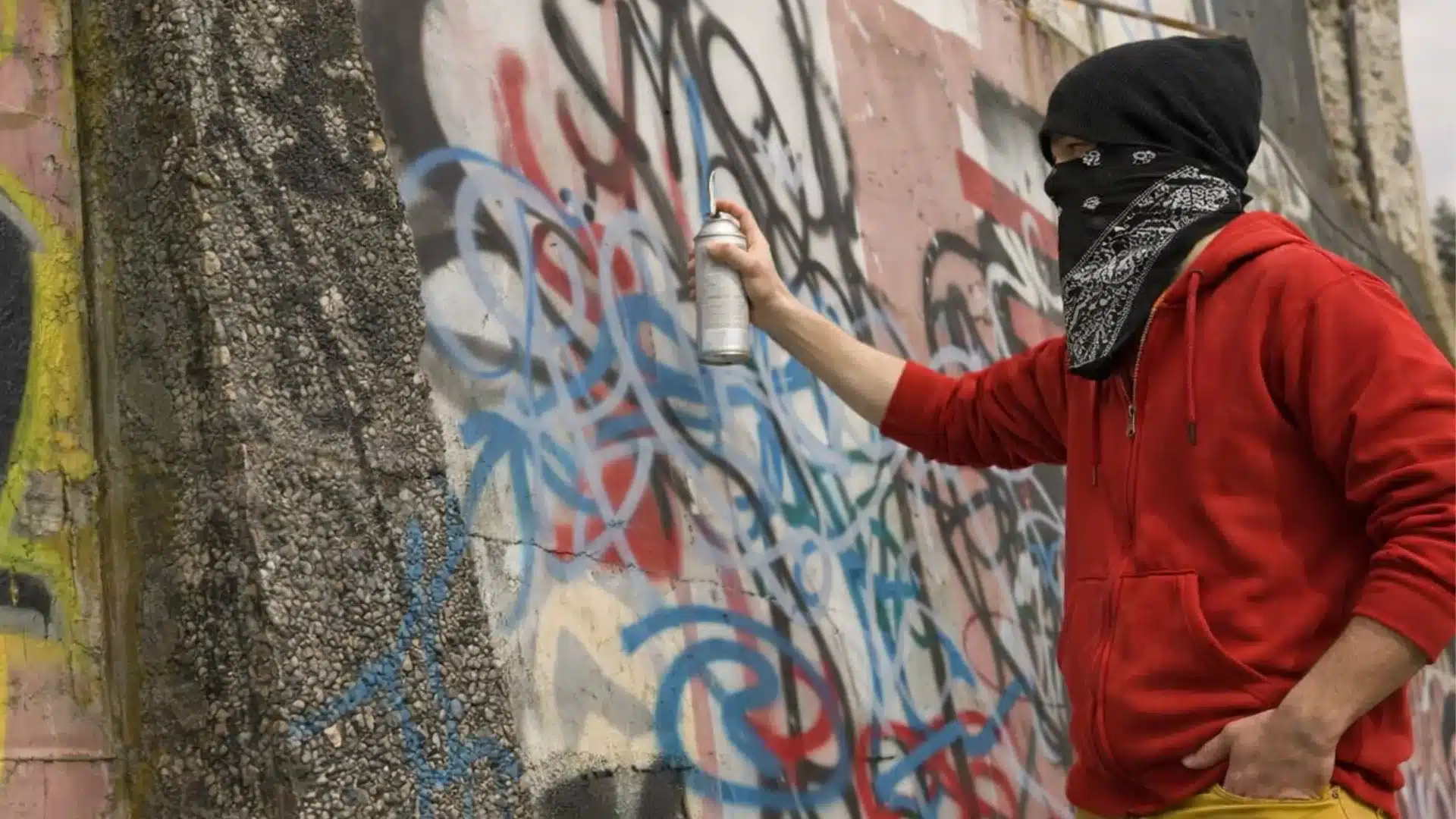 Masked street artist in red hoodie spray painting colorful graffiti on a rough concrete wall in an urban setting