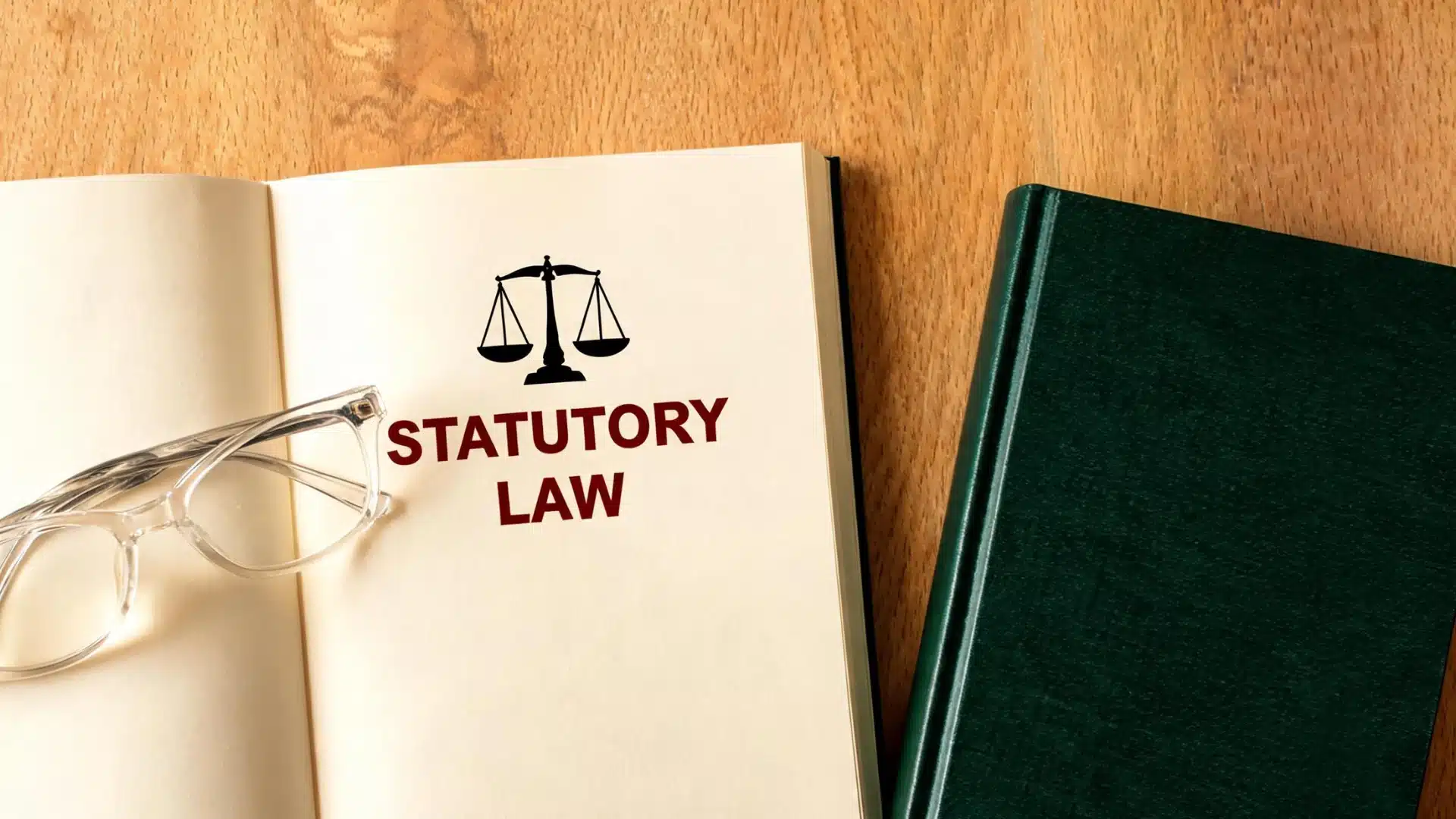 Open book with statutory law text, scales icon, eyeglasses, and green legal book on wooden desk