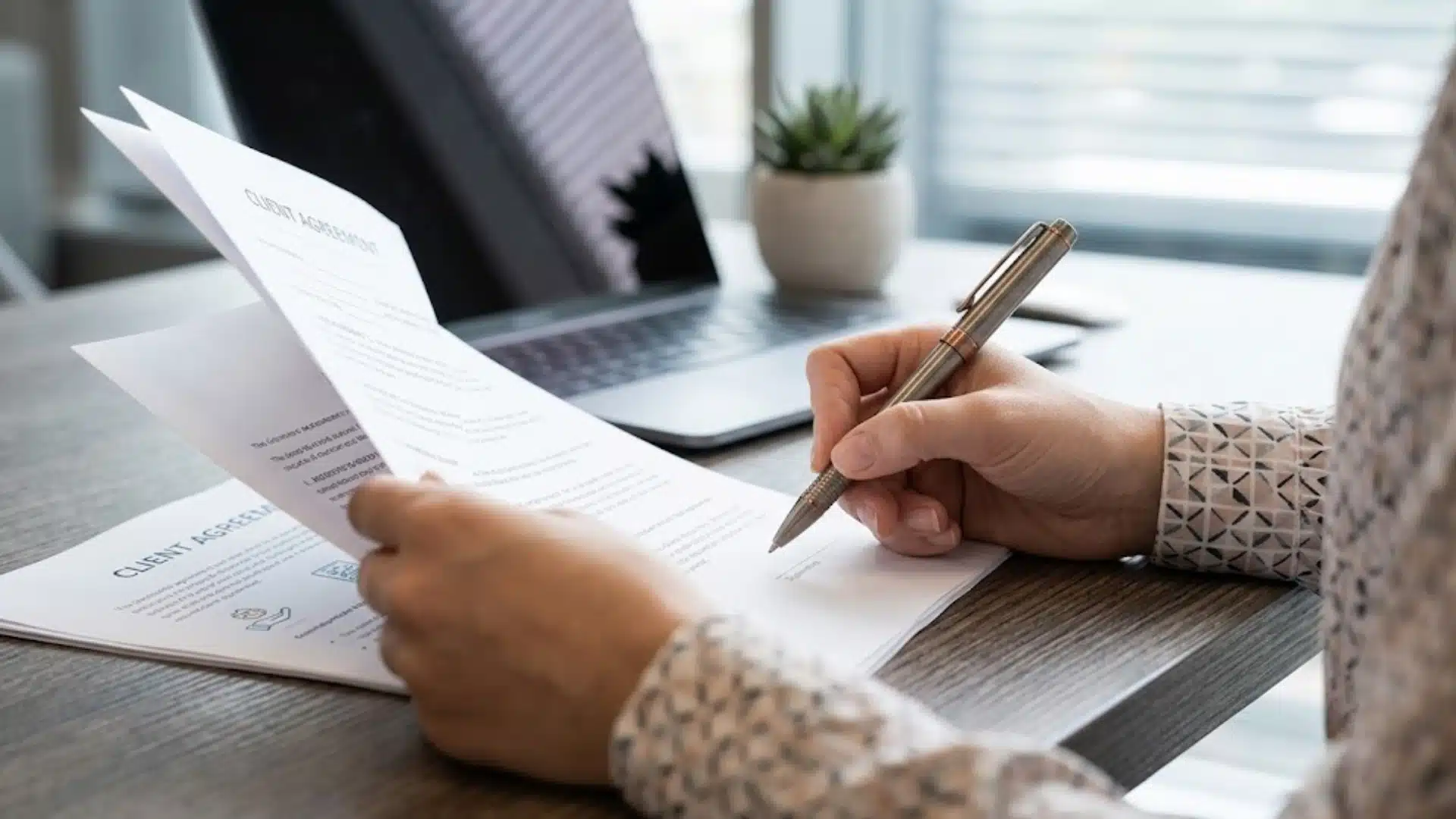 Person reviewing and signing a client agreement at a desk with laptop, papers, pen, and small office plant