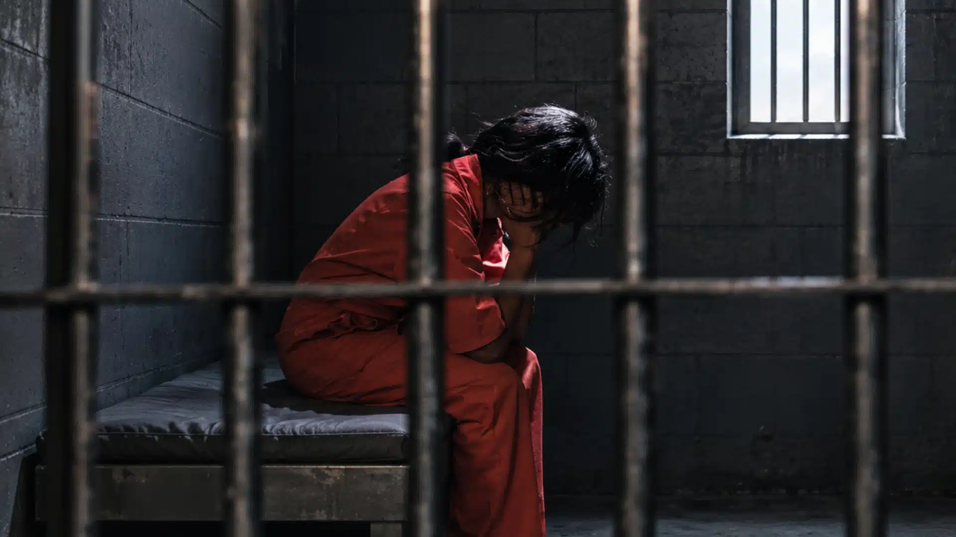 Person sitting in a dim prison cell behind bars with concrete walls and dramatic lighting