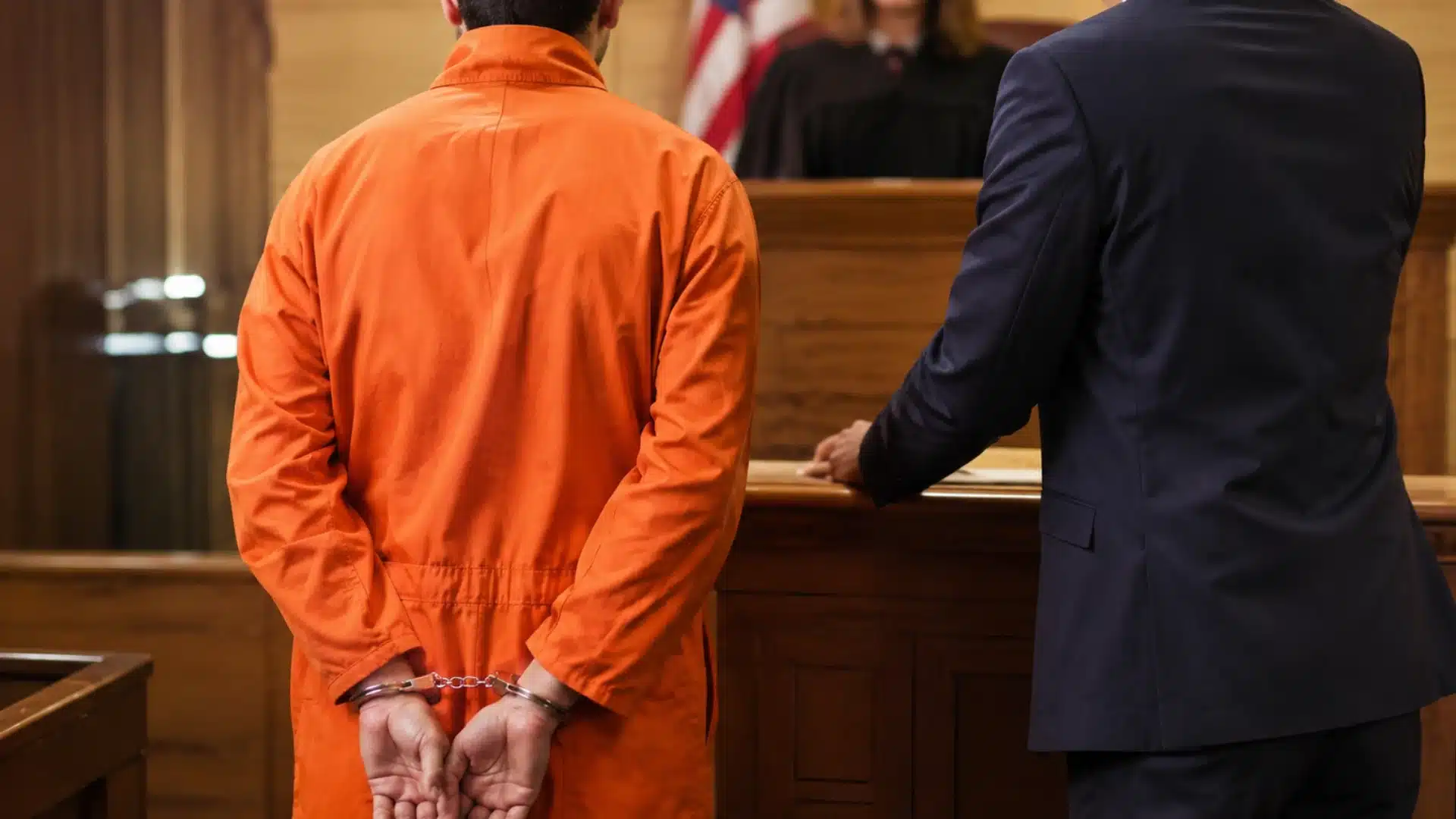 Handcuffed defendant and attorney stand before a judge in a wood-paneled courtroom